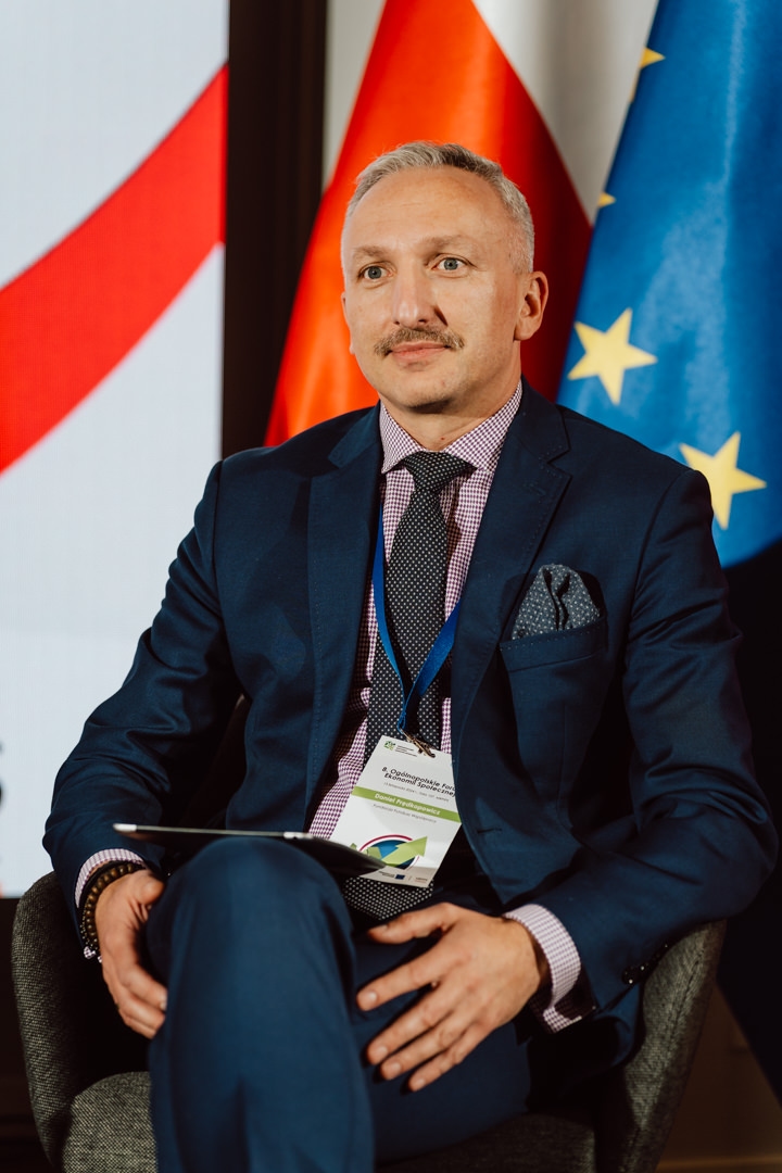 Ein Mann in einem marineblauen Anzug mit einem Schlüsselband und einem Konferenzabzeichen sitzt auf einem Stuhl. Hinter ihm sind verschwommene rote, weiße und blaue Flaggen mit gelben Sternen zu sehen, die auf eine internationale oder offizielle Veranstaltung hindeuten. 