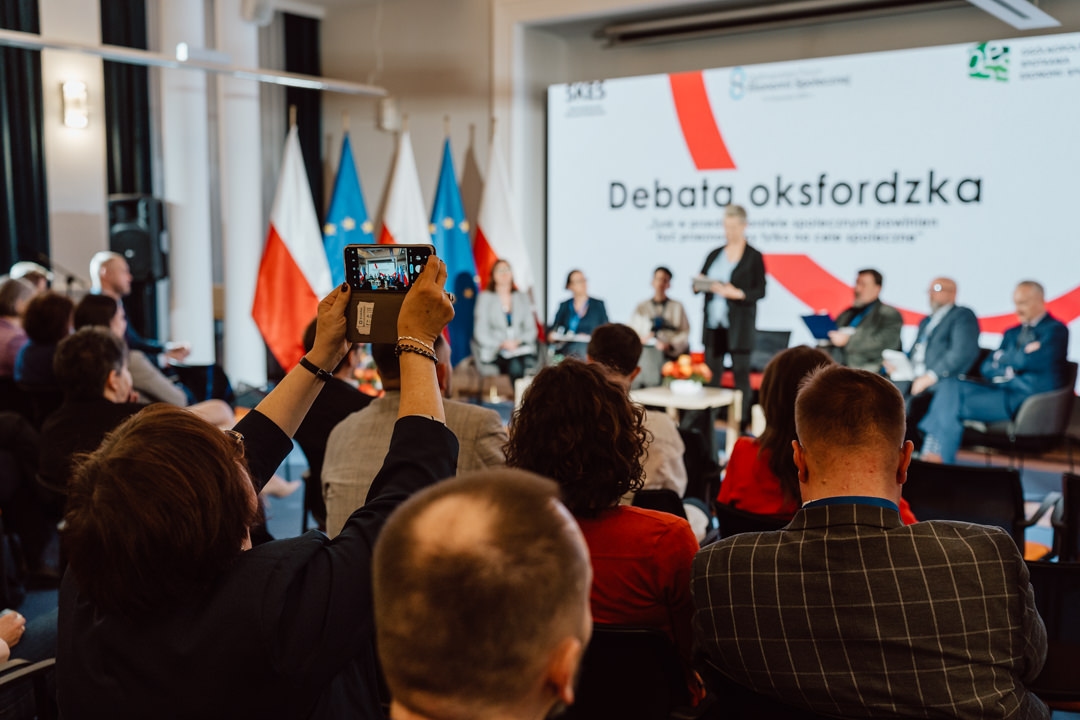 Ein Publikum verfolgt eine Podiumsdiskussion auf einer Bühne, auf der mehrere Redner sitzen; jemand im Vordergrund nimmt ein Telefon in die Hand, um ein Foto zu machen. Im Hintergrund sind polnische Flaggen und ein großer Bildschirm zu sehen, auf dem die 'Oxford-Debatte' gezeigt wird. 