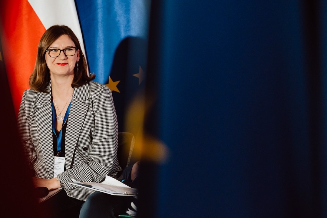 Eine Frau mit braunem Haar und Brille, die eine karierte Jacke und ein Schlüsselband trägt, sitzt lächelnd vor verschwommenen polnischen und EU-Flaggen. Sie hält Dokumente in der Hand und scheint an einer Veranstaltung oder Konferenz teilzunehmen. 