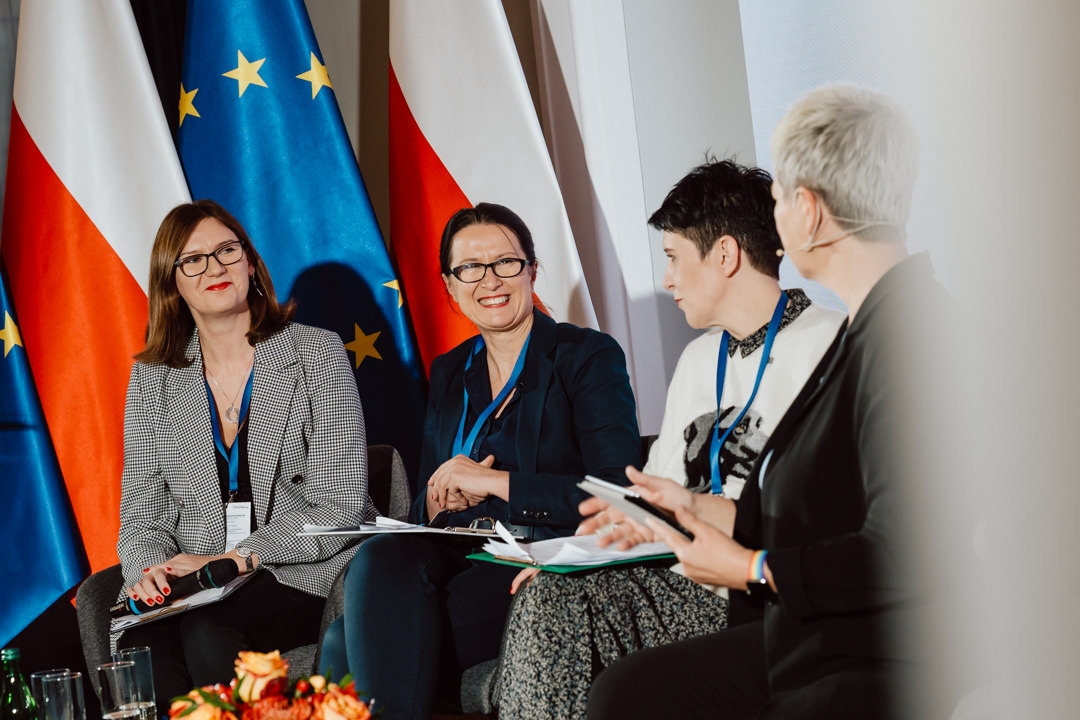 Vier Frauen sitzen und sprechen während einer Podiumsdiskussion, lächelnd und mit Dokumenten in der Hand. Hinter ihnen wehen die Flaggen von Polen und der Europäischen Union. Die Atmosphäre scheint professionell und kooperativ zu sein.  