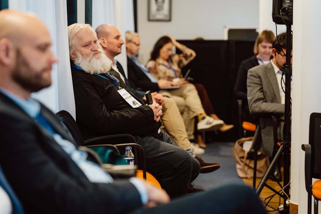 Eine Gruppe von Menschen sitzt auf Stühlen bei einer Konferenz oder einem Seminar. Die meisten von ihnen tragen Geschäftskleidung und Schlüsselbänder, einige hören aufmerksam zu und andere sehen entspannt aus. Die Atmosphäre wirkt professionell und engagiert.  