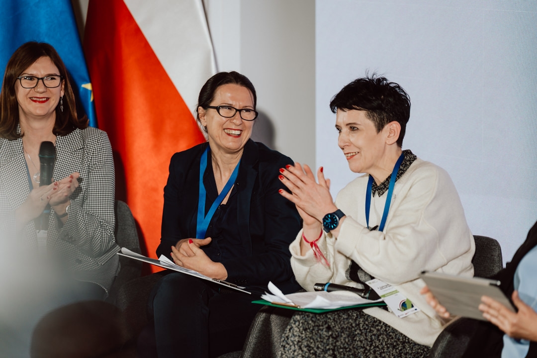 Drei Frauen sitzen während einer Podiumsdiskussion und lächeln. Zwei von ihnen halten Dokumente in der Hand und tragen Schlüsselbänder. Die Frau auf der linken Seite applaudiert und im Hintergrund sind Nationalflaggen zu sehen.  