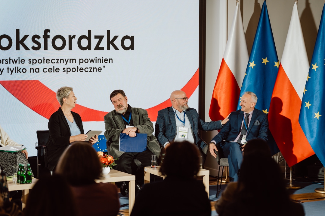 Vier Personen sitzen auf einer Bühne und diskutieren. Hinter ihnen sind die Flaggen Polens und der Europäischen Union zu sehen und der polnische Text wird auf eine große Leinwand projiziert. Im Vordergrund ist das Publikum zu sehen. 