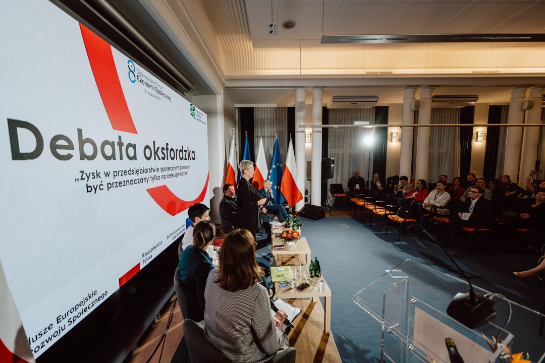 Ein Redner wendet sich während einer formellen Debatte in einem modernen Konferenzraum an das Publikum. Auf einem großen Bildschirm im Hintergrund wird der Titel der Veranstaltung auf Polnisch angezeigt. Die Diskussionsteilnehmer sitzen an einem Tisch; Fahnen und Mitglieder des Publikums sind zu sehen.  