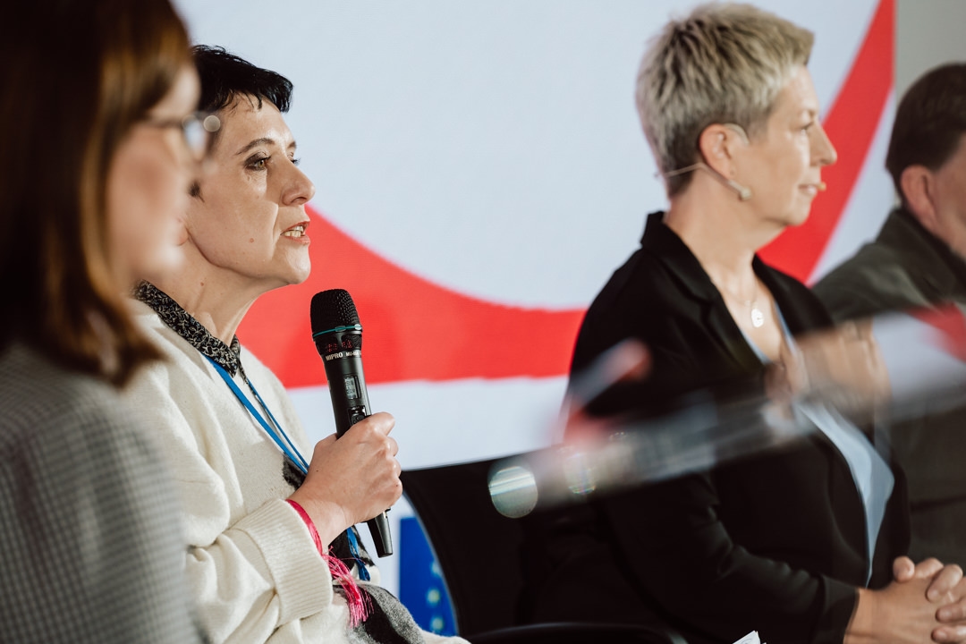 Eine Frau spricht in ein Mikrofon, während sie mit zwei anderen Personen auf einem Podium sitzt. Alle drei sind in Geschäftskleidung gekleidet und schauen konzentriert. Im Hintergrund ist ein großes, abstraktes rot-weißes Muster zu sehen.  