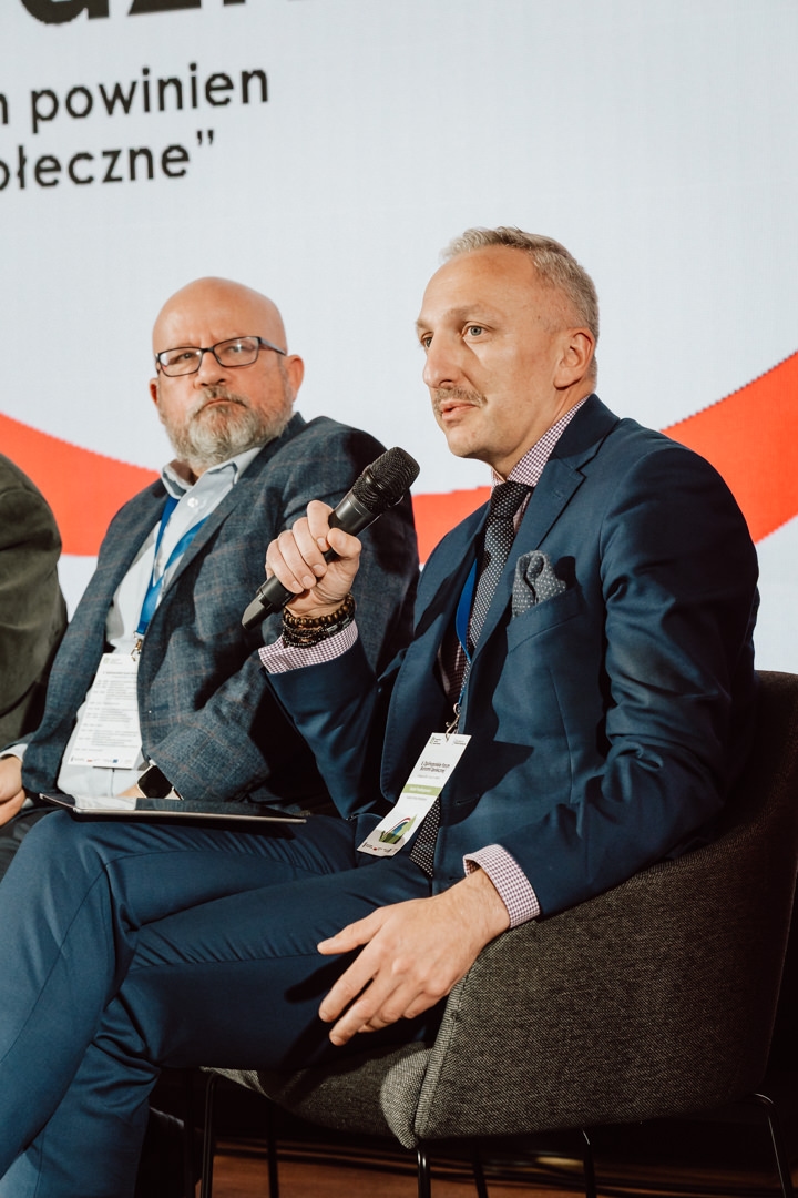 Zwei Männer in Anzügen sitzen während einer Podiumsdiskussion auf der Bühne. Einer von ihnen spricht in ein Mikrofon, während der andere aufmerksam zuhört. Beide tragen Konferenzschilder und hinter ihnen ist ein Präsentationsbildschirm mit Text zu sehen.  