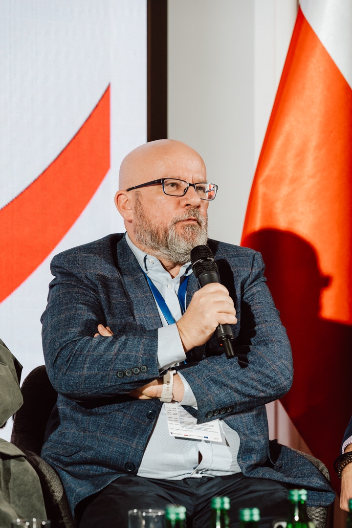 Ein glatzköpfiger Mann mit Brille, Bart und blauer Jacke hält ein Mikrofon, während er auf der Veranstaltung sitzt und spricht. Im Hintergrund ist eine große rot-weiße Flagge zu sehen. 