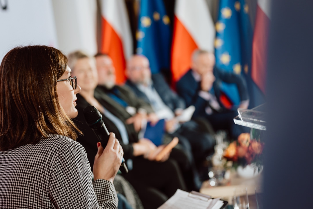 Eine Frau, die ein Mikrofon hält, spricht während einer Podiumsdiskussion. Im Hintergrund sitzen mehrere Personen in einer Reihe mit undeutlichen Flaggen Polens und der Europäischen Union hinter ihnen. 