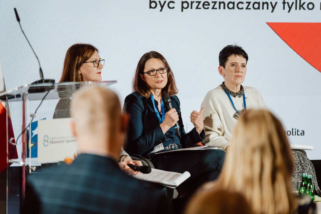 Drei Frauen sitzen auf der Bühne einer Podiumsdiskussion. Eine spricht in ein Mikrofon, während die anderen zuhören. Im Hintergrund ist ein Präsentationsbildschirm mit Text zu sehen. Im Vordergrund sind mehrere Zuhörer zu sehen.  