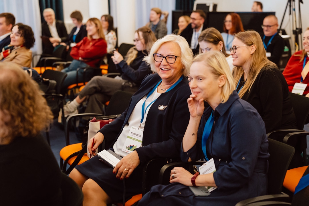 Eine Gruppe von Menschen sitzt in Reihen bei einer Konferenz oder einem Seminar. Zwei Frauen in der ersten Reihe lächeln und sprechen, während andere aufmerksam zuhören oder nach vorne schauen. Viele Teilnehmer tragen Anstecknadeln und Business-Kleidung.  