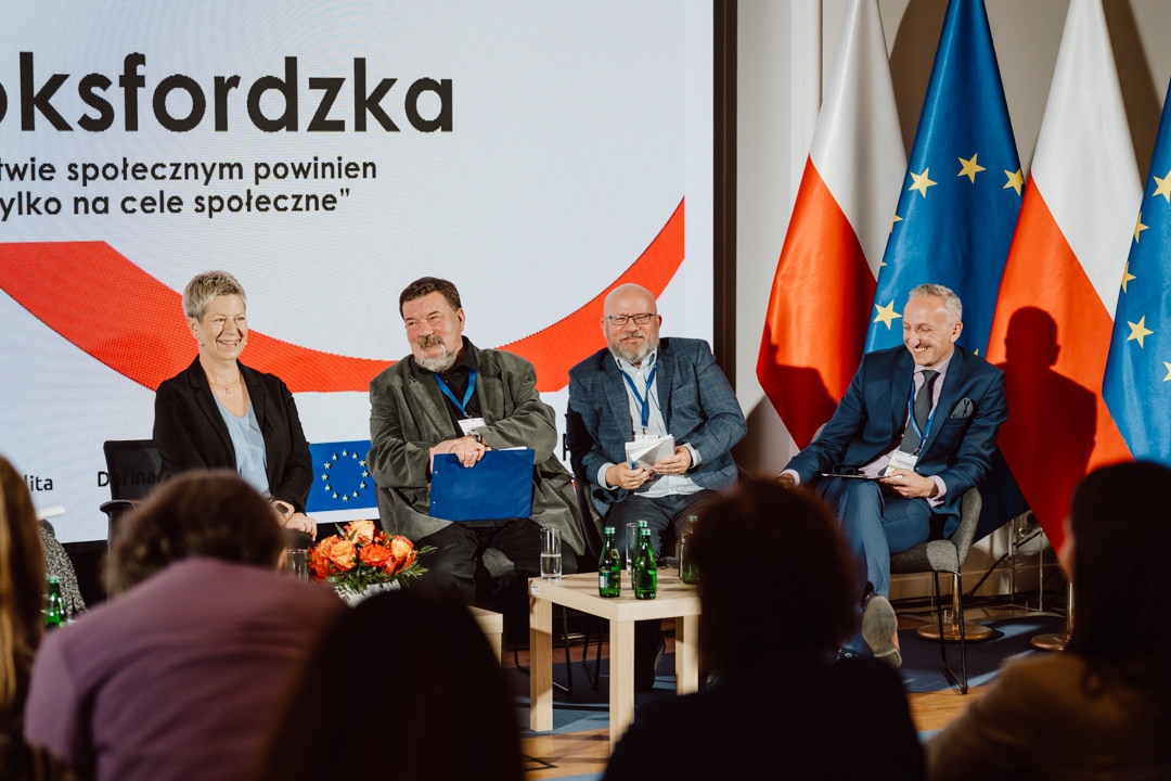 Vier Personen sitzen während einer Podiumsdiskussion auf der Bühne, das Publikum ist im Vordergrund zu sehen. Sie lächeln, hinter ihnen sind die Flaggen der Europäischen Union und Polens zu sehen, und auf einem großen Bildschirm wird polnischer Text angezeigt. 
