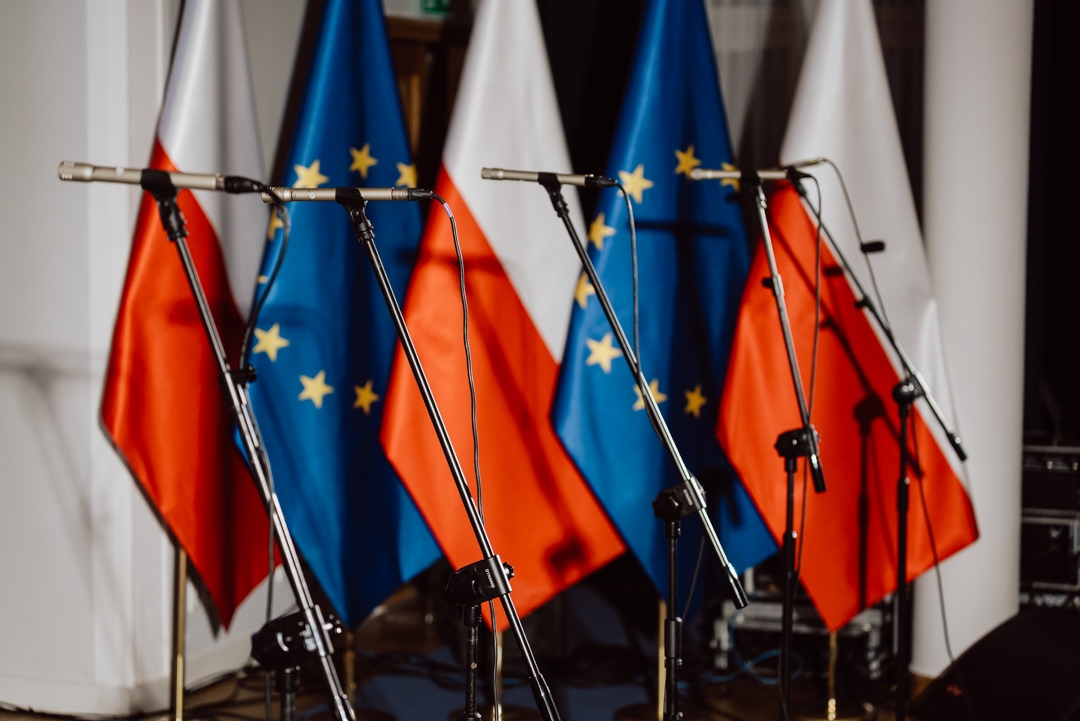 Mehrere Mikrofone sind vor den Flaggen Polens und der Europäischen Union aufgestellt. Das deutet auf eine Bühne hin, die für eine Rede, eine Konferenz oder eine offizielle Veranstaltung vorbereitet ist. Es sind keine Menschen auf dem Foto zu sehen. 