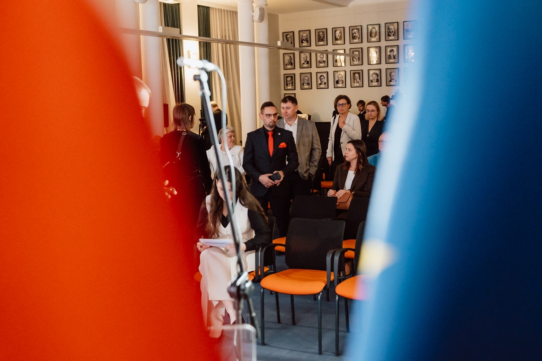 In einem Raum mit orangefarbenen Stühlen sind Menschen versammelt, einige stehen, andere sitzen. Das Foto wird von unscharfen blauen und orangefarbenen Formen im Vordergrund eingerahmt. An der Wand im Hintergrund sind Porträts aufgereiht. Ein Mikrofon ist ebenfalls zu sehen.   