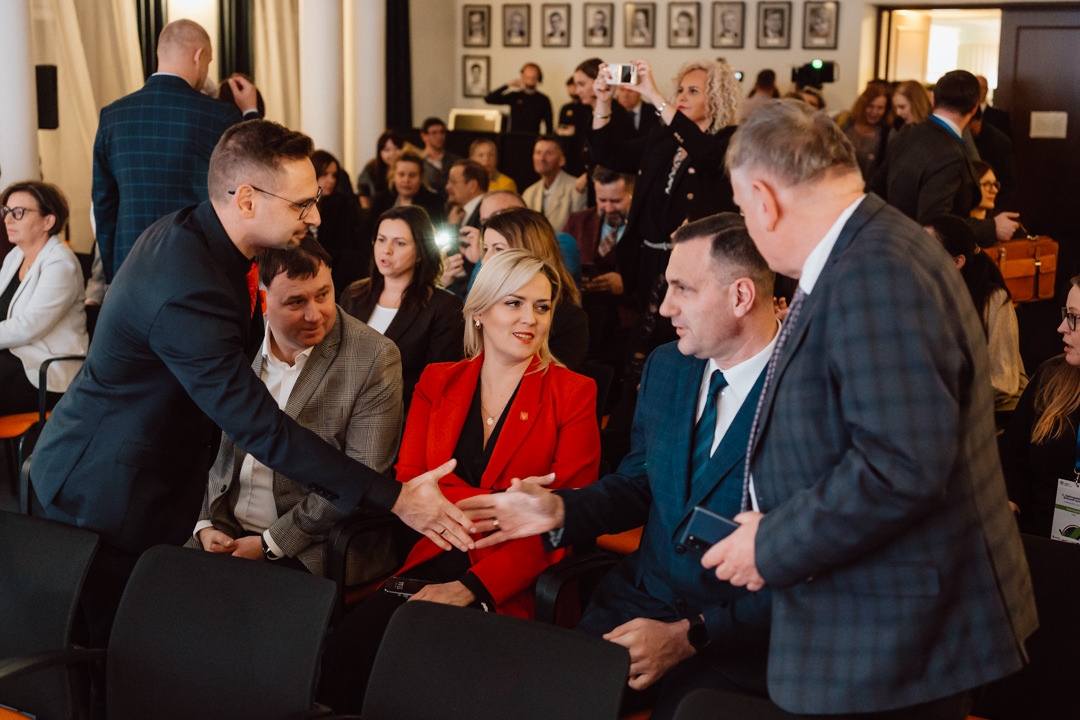 Eine Gruppe von Menschen in Geschäftskleidung interagiert in einem Konferenzraum. Zwei Männer im Vordergrund schütteln sich die Hände, während andere im Hintergrund zusehen, reden oder Fotos machen.