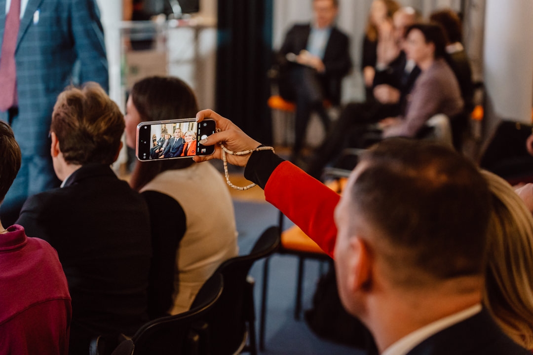 Eine Person in einer roten Jacke hält ein Smartphone in der Hand, um ein Foto oder ein Video während einer Veranstaltung in einem Raum zu machen, in dem mehrere Personen sitzen und den Redner im Hintergrund beobachten.
