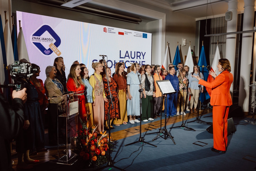 Ein Chor von Männern und Frauen steht auf der Bühne und singt, angeführt von einer Frau in einem orangefarbenen Anzug. Fahnen und ein Blumenstrauß sind zu sehen, während hinter ihnen ein großer Bildschirm die 'Social Laurels' und das Logo der Veranstaltung anzeigt. 