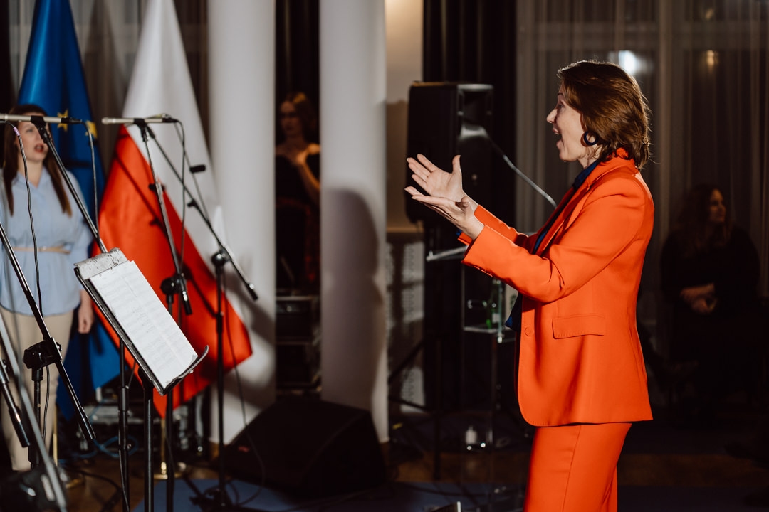 Eine Frau in einem orangefarbenen Overall dirigiert oder spricht leidenschaftlich vor Mikrofonen und Notenständern, während im Hintergrund polnische Flaggen und mehrere Menschen zu sehen sind.