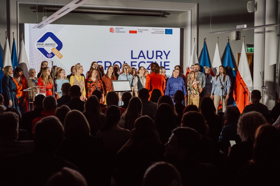 Eine Gruppe von Menschen steht gemeinsam auf der Bühne vor einem großen Bildschirm, auf dem Text bei einer Veranstaltung in einer Halle angezeigt wird, während das Publikum sitzt und zusieht. Rechts von der Bühne sind Fahnen zu sehen. 
