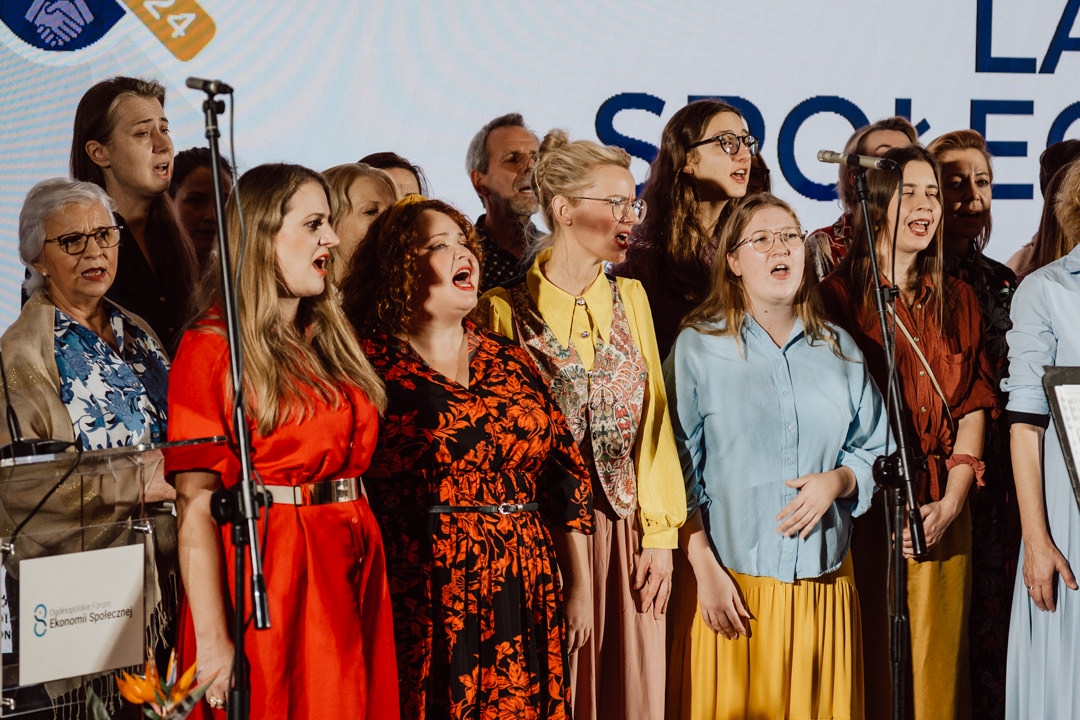 Eine bunt gemischte Gruppe von Menschen in farbenfroher, halb-formeller Kleidung steht zusammen auf der Bühne und singt in Mikrofone. Ein Bildschirm hinter ihnen zeigt einige der Logos und Texte an. 