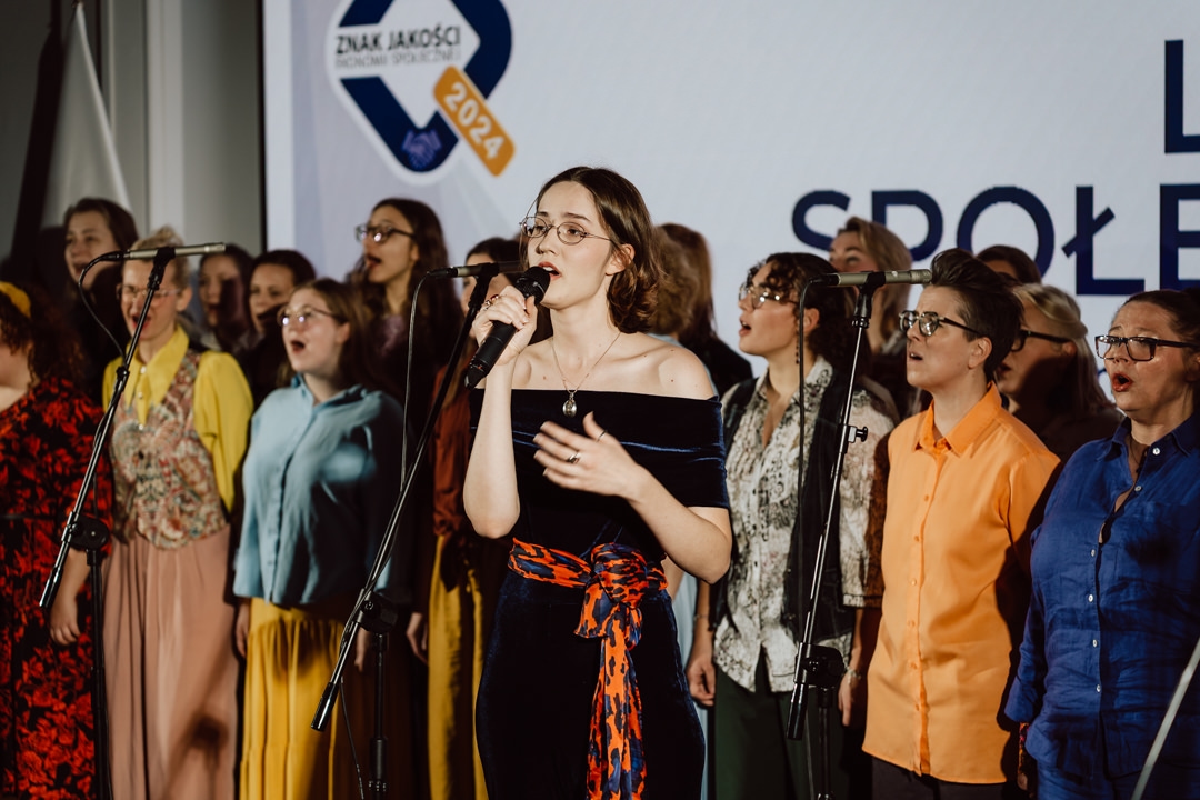 Eine Frau singt auf der Bühne in ein Mikrofon, umgeben von einem bunten Chor. Die Gruppe steht vor einem großen Bildschirm, auf dem die Worte 'QUALITY SIGN 2024' mit blauen und gelben Grafiken zu sehen sind. 