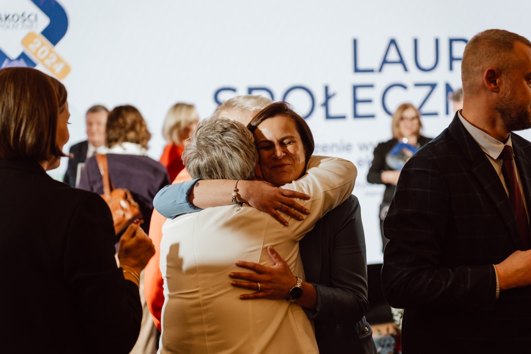 Zwei Frauen umarmen sich herzlich im Vordergrund während einer Zimmerparty, während andere Leute im Hintergrund stehen und sich unterhalten. Hinter ihnen ist ein großes, teilweise sichtbares Schild zu sehen. 