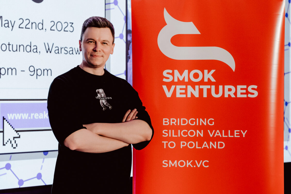 fotograf startupow rotunda reaktorX 1 Ein Mann steht lächelnd mit verschränkten Armen neben einem roten SMOK Ventures-Banner mit der Aufschrift 'Bridging Silicon Valley to Poland'. Auf einem Bildschirm im Hintergrund werden Details zur Veranstaltung angezeigt, darunter das Datum und der Veranstaltungsort in Warschau.