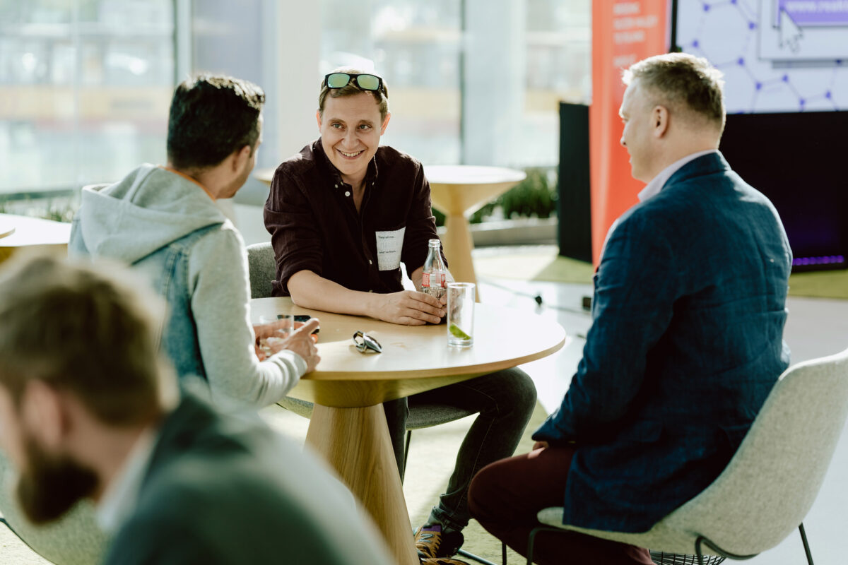 fotograf startupow rotunda reaktorX 12 Drei Personen sitzen um einen Tisch in einem hellen, modernen Raum und unterhalten sich. Eine Person in der Mitte lächelt und hält eine Flasche, während die anderen aufmerksam zuhören. Die vierte Person befindet sich unscharf im Vordergrund.