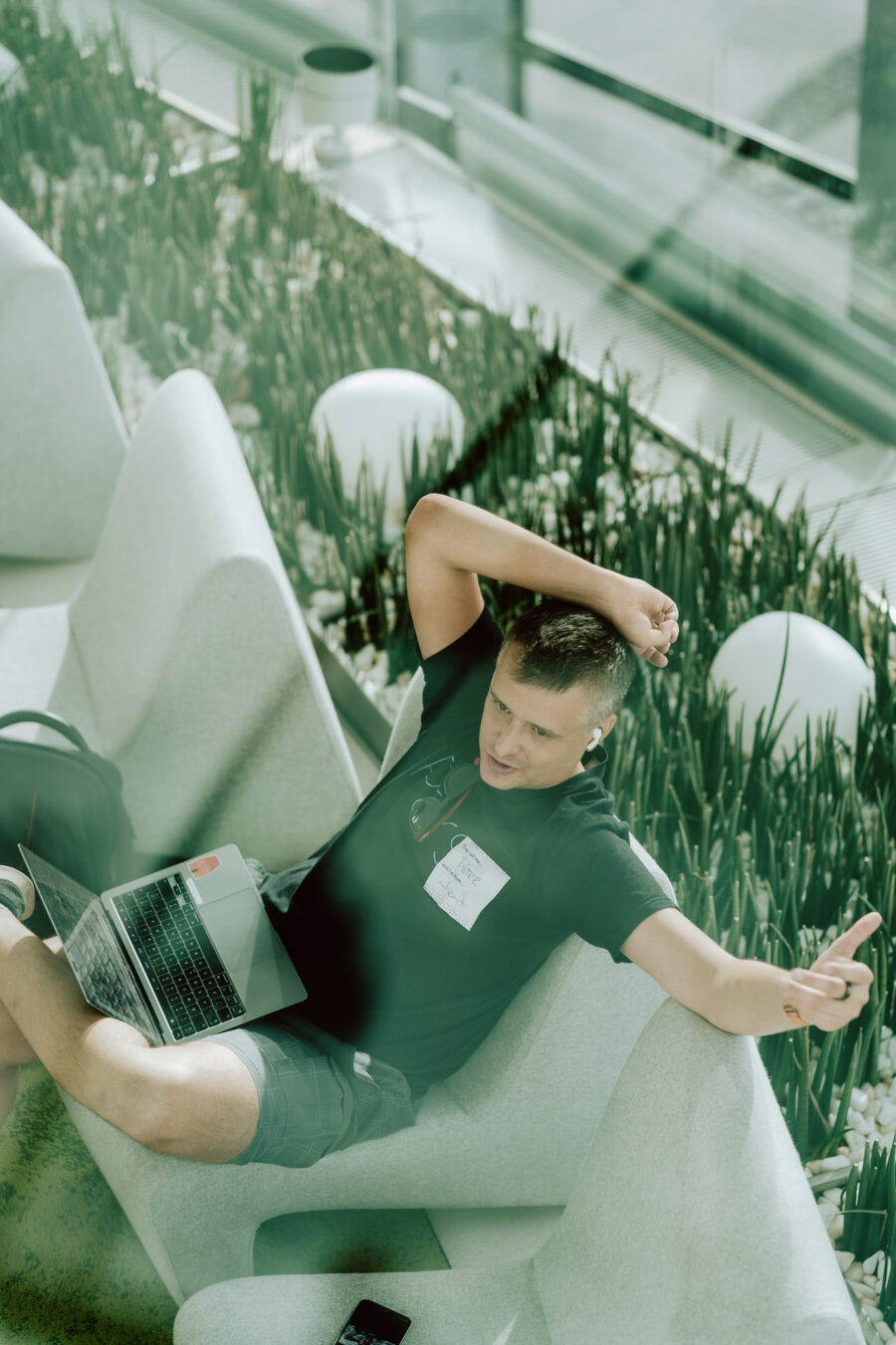 fotograf startupow rotunda reaktorX 14 Eine Person sitzt lässig auf einem hellen Sofa mit einem Laptop auf dem Schoß, bekleidet mit einem schwarzen T-Shirt und Shorts, eine Hand hinter dem Kopf verschränkt, in einem hellen, modernen Interieur mit Pflanzen.