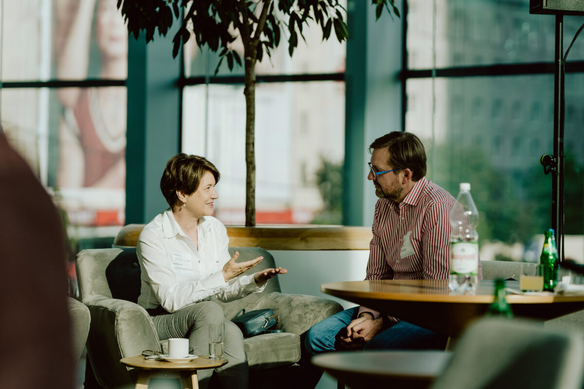 fotograf startupow rotunda reaktorX 15 Zwei Personen sitzen in Sesseln in einem modernen, sonnendurchfluteten Wohnzimmer und führen ein Gespräch. Eine von ihnen gestikuliert mit ihren Händen, während die andere zuhört. Im Vordergrund steht ein Tisch mit Wasserflaschen und Tassen.