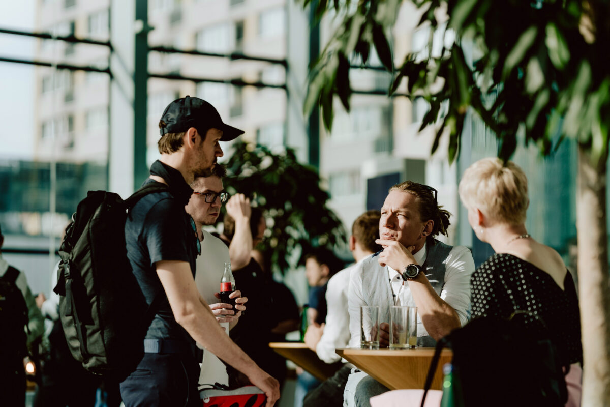 fotograf startupow rotunda reaktorX 16 Eine Gruppe von Leuten trifft sich an einem hohen Tisch in einem sonnenbeschienenen Innenraum mit großen Fenstern und Pflanzen, Getränke in der Hand und mit Blick auf die Stadt draußen.
