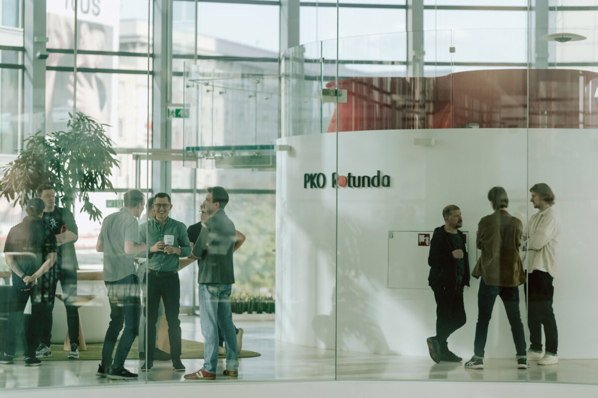 fotograf startupow rotunda reaktorX 17 Eine Gruppe von Menschen steht und unterhält sich in einem modernen Gebäude mit Glaswänden in der Nähe des Schildes "PKO Rotunda". Einige halten Getränke in der Hand und unterhalten sich in kleinen Gruppen, während das Sonnenlicht hereinströmt.