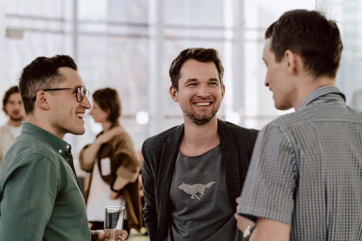 fotograf startupow rotunda reaktorX 19 Drei Männer stehen zusammen in einem Raum, lächeln und unterhalten sich. Einer von ihnen hält ein Glas Wasser in der Hand und ein anderer trägt eine Jacke über einem T-Shirt mit Tiermotiven. Im Hintergrund sehen Sie andere Menschen und große Fenster.