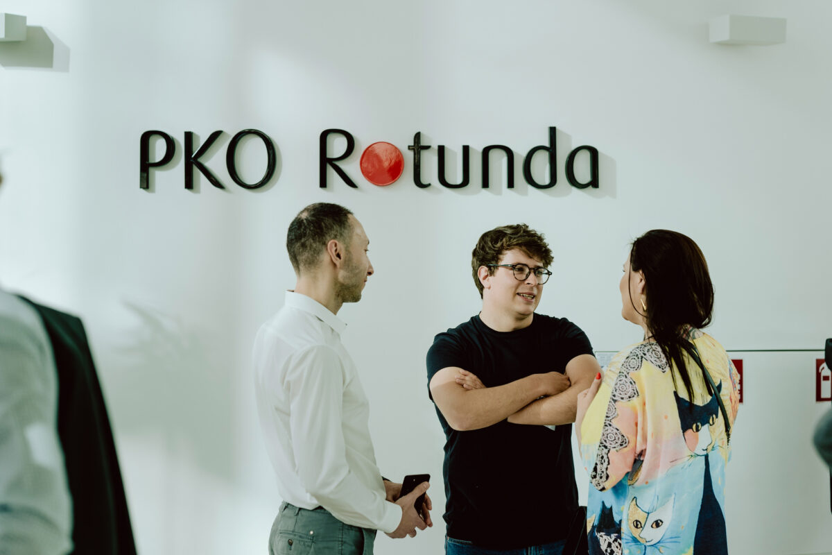 fotograf startupow rotunda reaktorX 2 Drei Personen stehen und unterhalten sich vor einer Wand mit dem Schild 'PKO Rotunda'. Der Mann links trägt ein weißes Hemd, der Mann in der Mitte trägt eine Brille und ein schwarzes Hemd und die Frau hat ein farbiges Oberteil mit einem Katzenaufdruck.