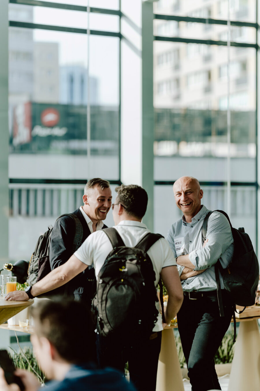 fotograf startupow rotunda reaktorX 21 Drei Männer mit Rucksäcken stehen und unterhalten sich lächelnd und lachend in einem hellen, modernen Raum mit großen Fenstern und den Gebäuden der Stadt draußen. Im Hintergrund sind andere Menschen und Tische zu sehen.