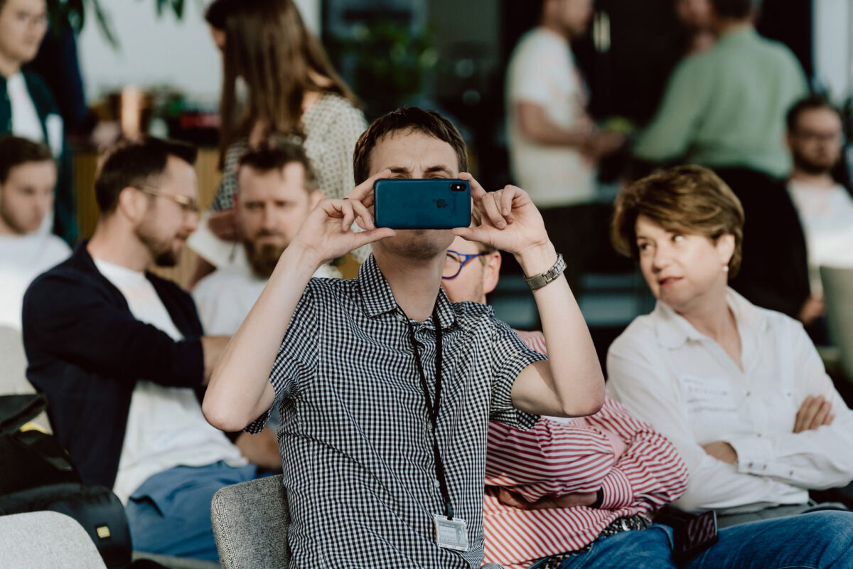 fotograf startupow rotunda reaktorX 22 Die Person im Vordergrund hält ein Smartphone in der Hand, um ein Foto oder ein Video aufzunehmen, während im Hintergrund mehrere Personen bei einer scheinbar zwanglosen Veranstaltung oder Besprechung im Raum sitzen und sich unterhalten.