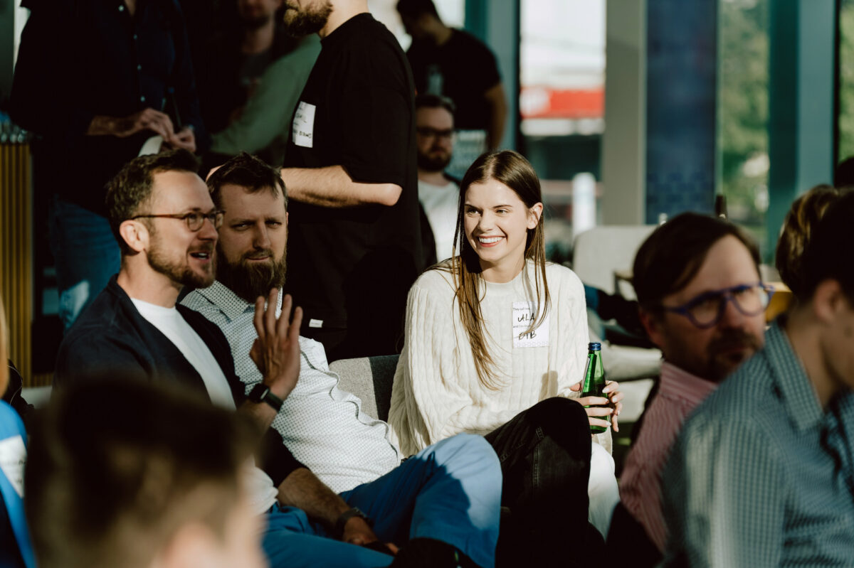fotograf startupow rotunda reaktorX 23 Eine Gruppe von Menschen sitzt während einer Party in einem Raum und unterhält sich. Eine Frau in einem weißen Pullover lächelt und hält eine Flasche, während andere sich unterhalten. Sonnenlicht strömt durch die großen Fenster im Hintergrund herein.