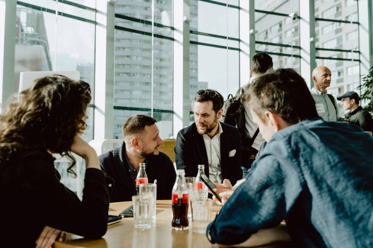 fotograf startupow rotunda reaktorX 24 Vier Personen sitzen an einem Tisch mit Getränken, unterhalten sich und lächeln in einem modernen, sonnenbeschienenen Raum mit großen Fenstern und städtischen Gebäuden im Hintergrund. Andere Personen sind im Hintergrund zu sehen.