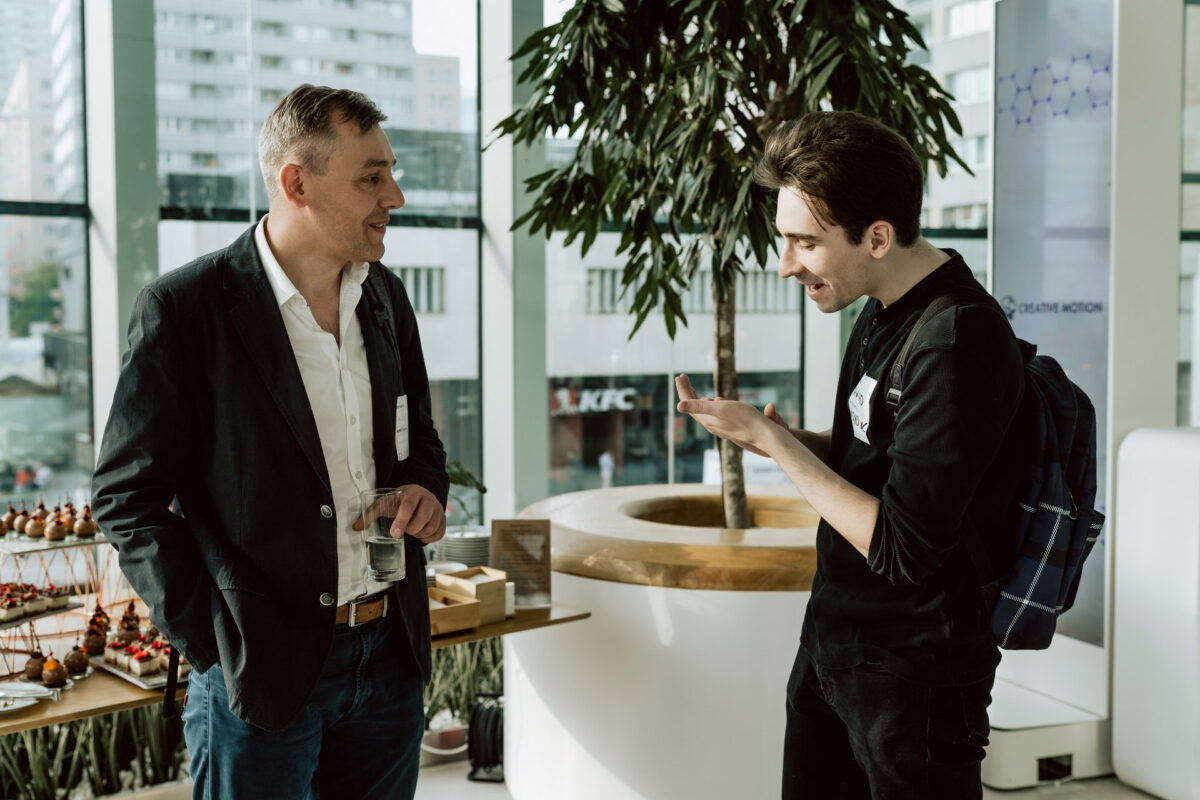 fotograf startupow rotunda reaktorX 25 Zwei Männer stehen in einem Raum bei einem gesellschaftlichen Ereignis, unterhalten sich und lächeln. Einer hält ein Getränk in der Hand, der andere schaut auf sein Telefon. Auf einem Tisch in der Nähe stehen Snacks, und im Hintergrund sind große Fenster mit städtischen Gebäuden zu sehen.