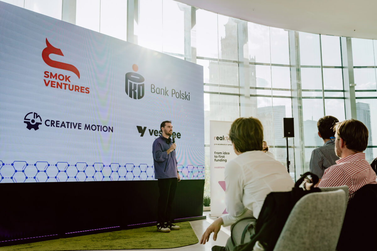 fotograf startupow rotunda reaktorX 27 Ein Mann, der auf der Konferenz spricht, steht auf der Bühne, während hinter ihm die Logos von Smok Ventures, Polski Bank, Creative Motion und Vestbee auf einem großen Bildschirm zu sehen sind. Mehrere sitzende Teilnehmer hören in dem modernen, sonnendurchfluteten Raum zu.