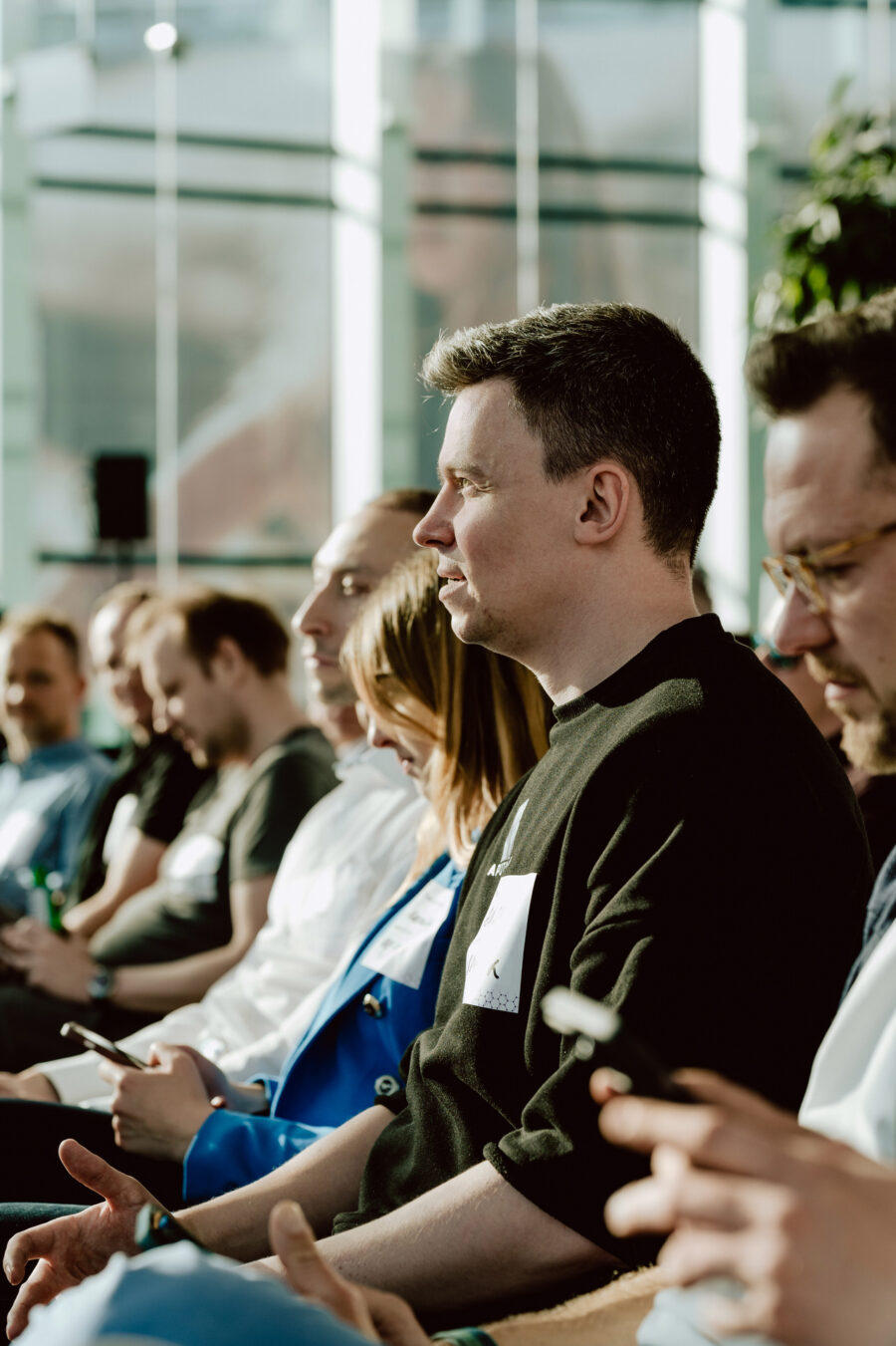 fotograf startupow rotunda reaktorX 28 Eine Gruppe von Menschen sitzt in einer Reihe in einem Raum, einige benutzen ihre Telefone, während das Sonnenlicht durch die großen Fenster hinter ihnen hereinströmt. Im Mittelpunkt der Aufmerksamkeit steht ein Mann in einem schwarzen Pullover, der aufmerksam vor sich hin starrt.