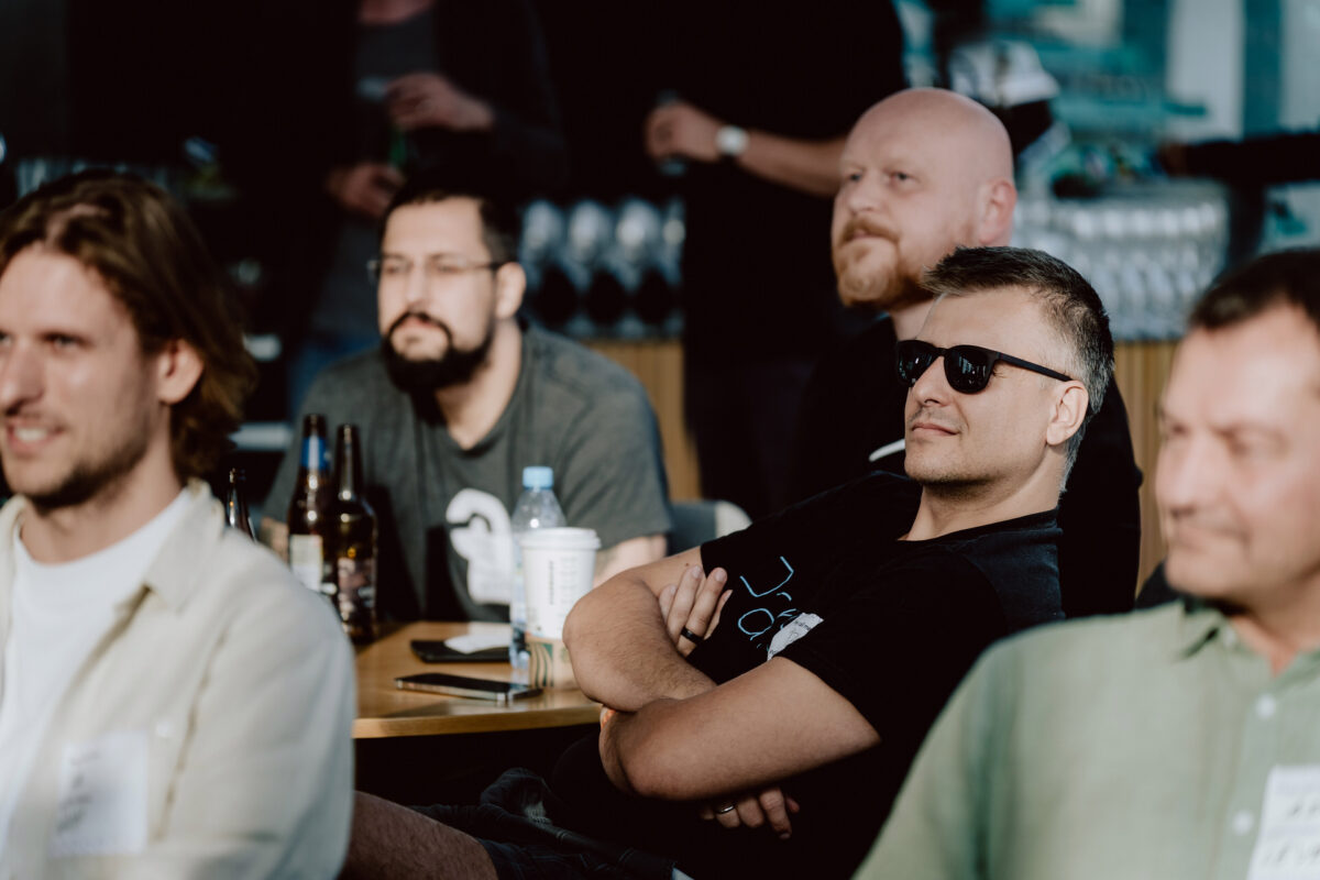 fotograf startupow rotunda reaktorX 29 Eine Gruppe von Männern sitzt an Tischen und beobachtet aufmerksam etwas im Hintergrund. Einer der Männer in der Mitte trägt eine Sonnenbrille und hat die Arme verschränkt. Auf den Tischen sind Getränke und Flaschen zu sehen, was auf eine zwanglose Veranstaltung oder ein Treffen hindeutet.