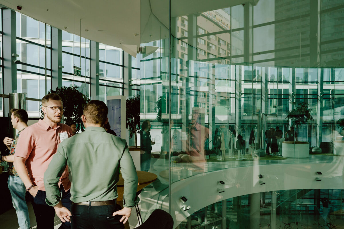 fotograf startupow rotunda reaktorX 3 Zwei Männer stehen und unterhalten sich in einem modernen, sonnendurchfluteten Glasgebäude mit Spiegelungen an den Wänden. Im Hintergrund stehen andere Menschen und Topfpflanzen, die eine lebendige, professionelle Atmosphäre schaffen.