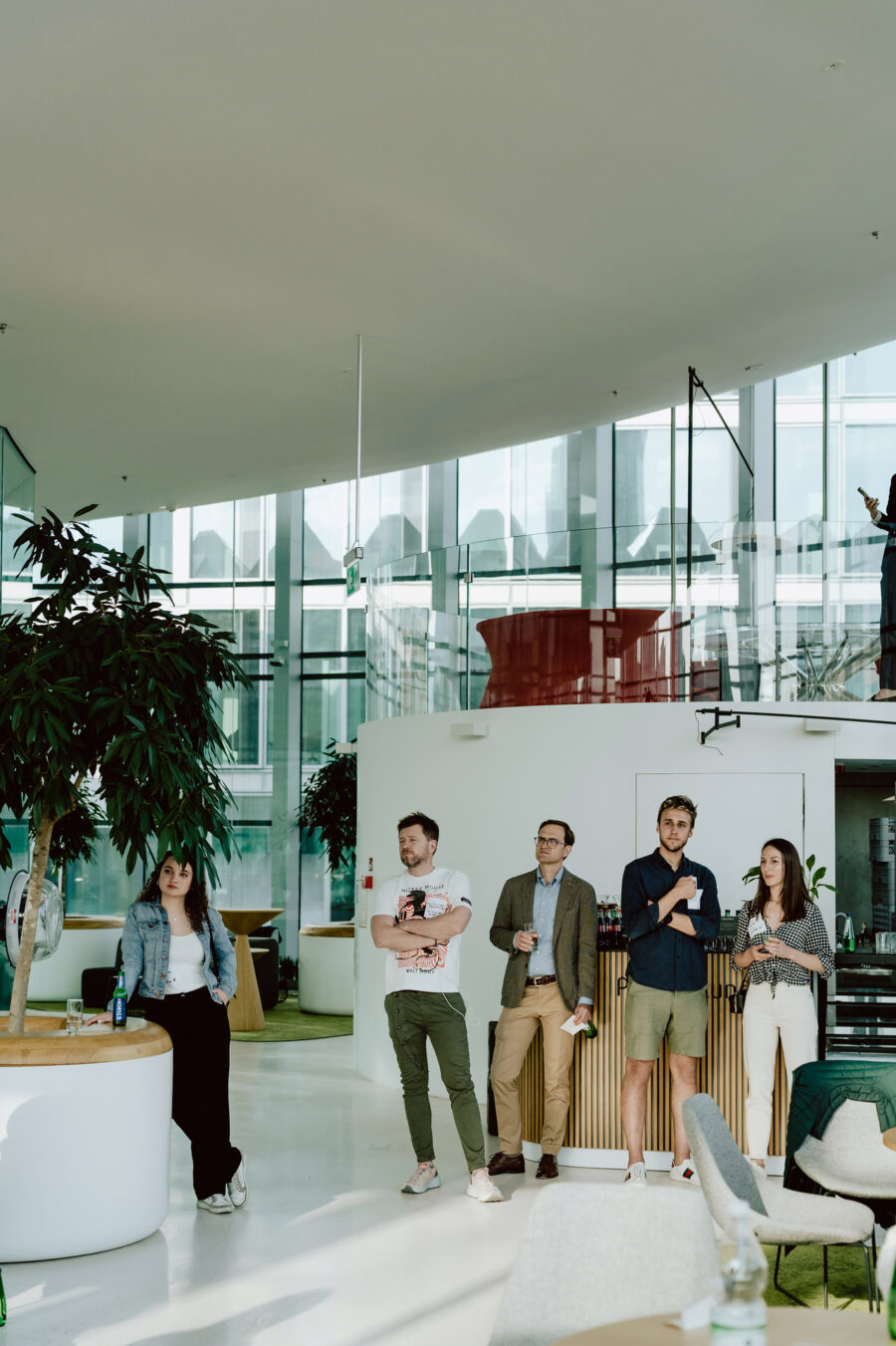 fotograf startupow rotunda reaktorX 30 Vier Personen stehen in einem Raum in der Nähe von großen Fenstern und Pflanzen und beobachten aufmerksam etwas im Hintergrund. Eine weitere Person sitzt auf einer geschwungenen weißen Bank auf der linken Seite. Der Raum ist modern und hat viel natürliches Licht.