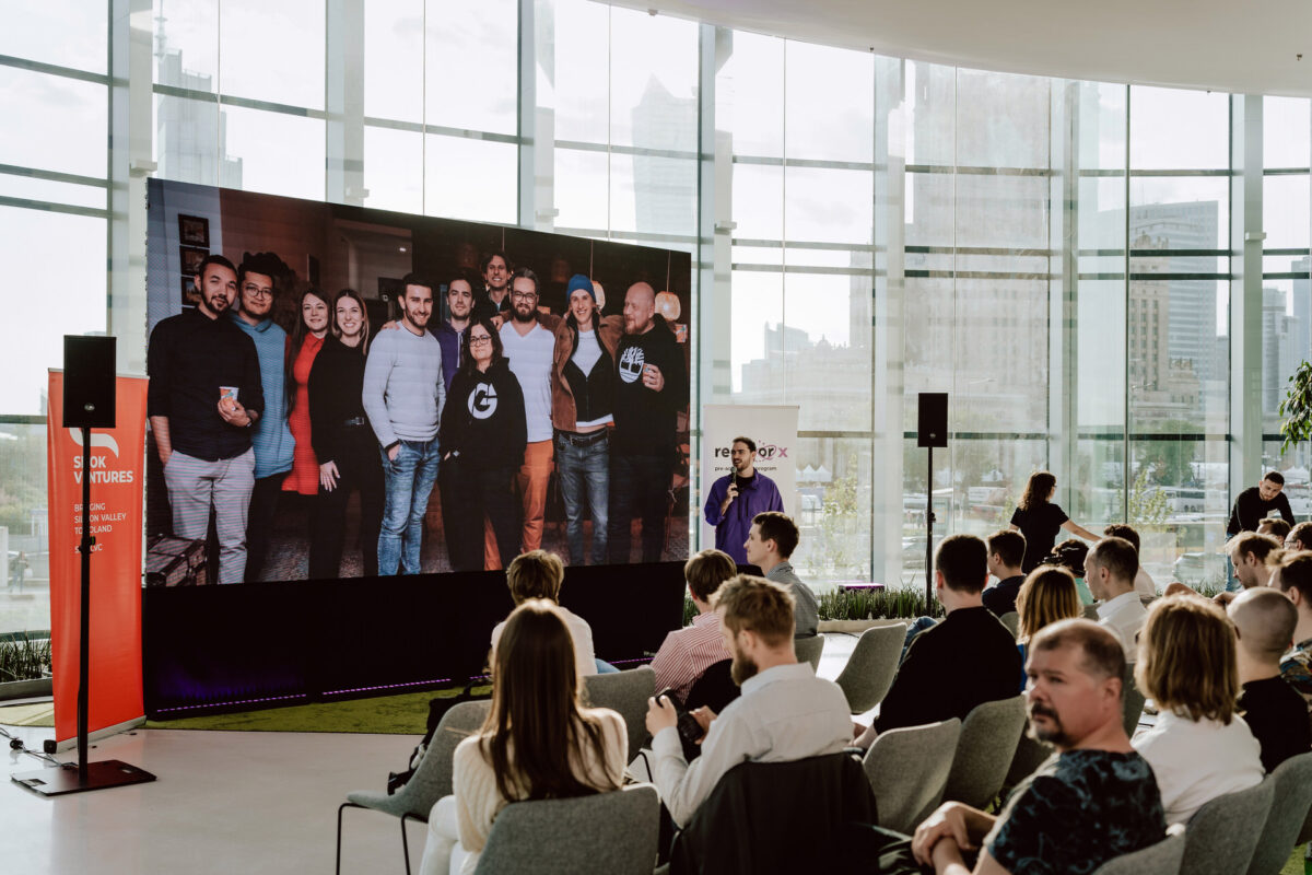 fotograf startupow rotunda reaktorX 31 Die Zuschauer sitzen in einem modernen Gebäude mit Glaswänden und sehen auf einen großen Bildschirm, der ein Gruppenfoto lächelnder Menschen zeigt, die zusammen in einem Raum stehen. Außerhalb der Fenster sind Gebäude der Stadt zu sehen.
