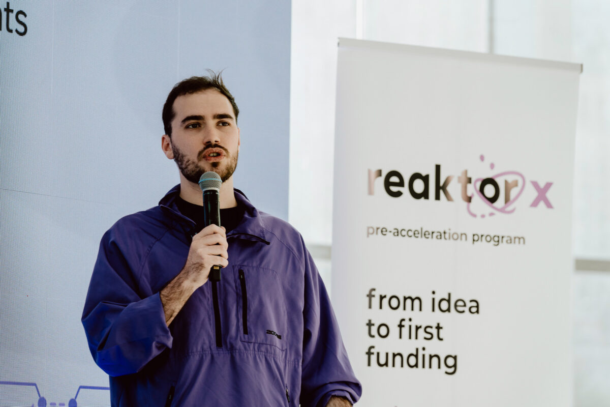 fotograf startupow rotunda reaktorX 32 Ein Mann in einer lila Jacke spricht in ein Mikrofon vor einem Banner mit der Aufschrift 'Reaktorx Vorbeschleunigungsprogramm' und den Worten 'von der Idee zur ersten Finanzierung'.