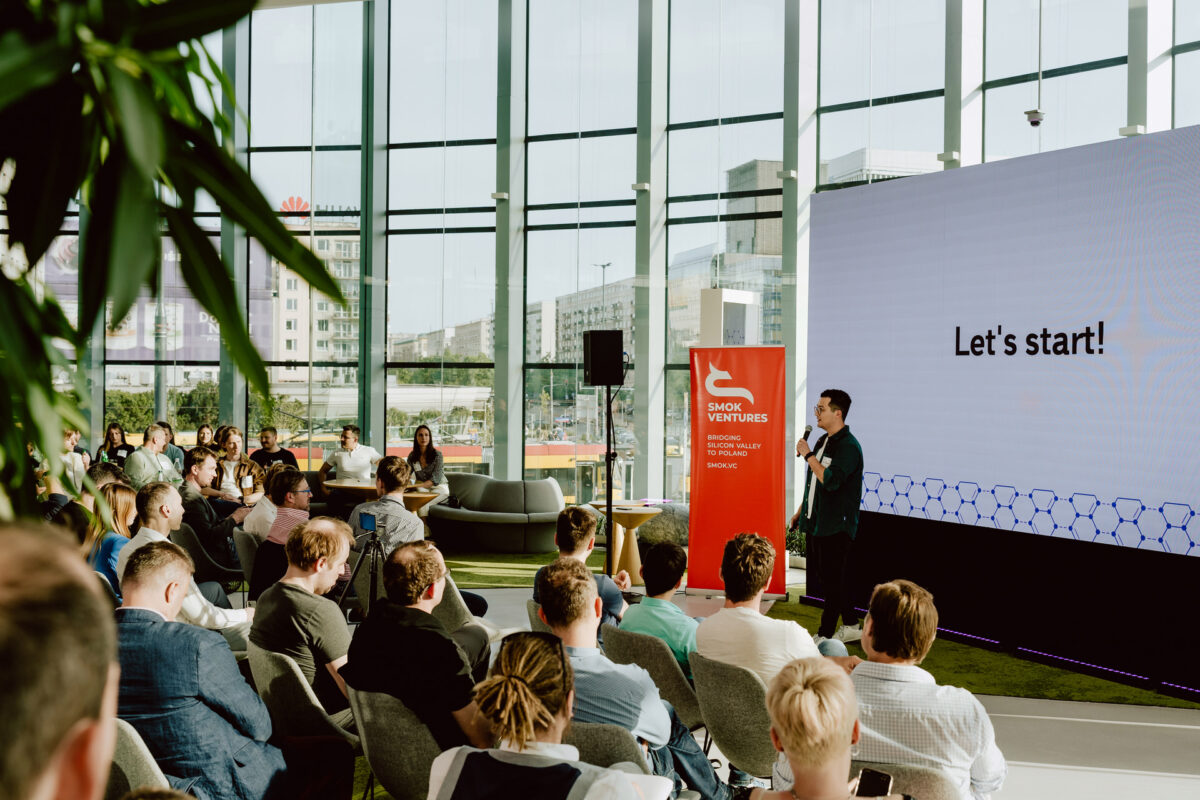 fotograf startupow rotunda reaktorX 33 Der Redner spricht zu einem sitzenden Publikum in einem hellen, modernen Raum mit großen Fenstern. Auf dem Bildschirm neben ihm stehen die Worte "Let's get started!" und ein rotes Schild verkündet "SMOK Ventures". Die Leute hören aufmerksam zu.
