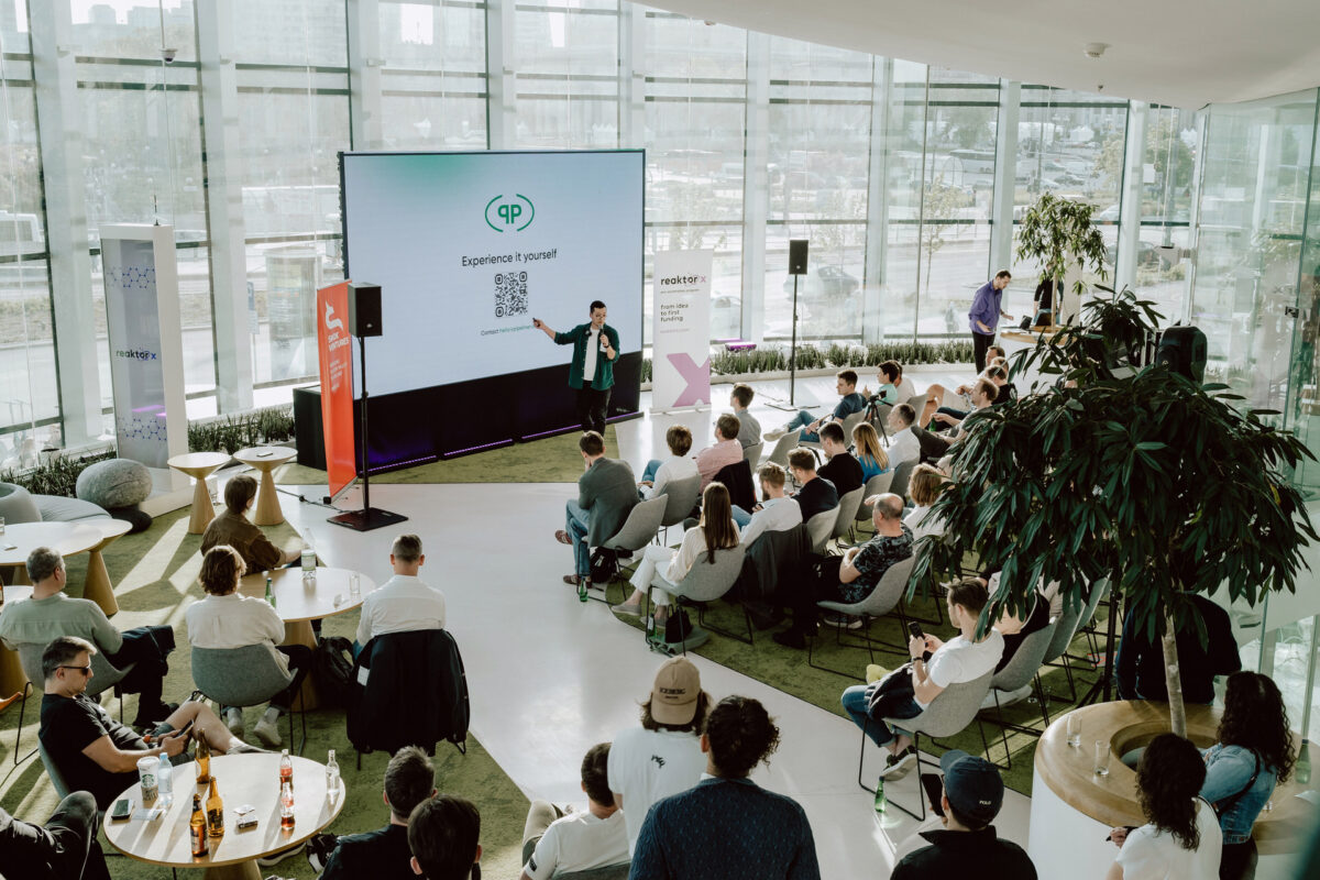 fotograf startupow rotunda reaktorX 34 Der Redner spricht zu den Zuhörern in einem modernen, gläsernen Konferenzraum. Die Leute sitzen an Tischen und in Reihen, hören zu und schauen auf den großen Bildschirm an der Vorderseite des Raums.