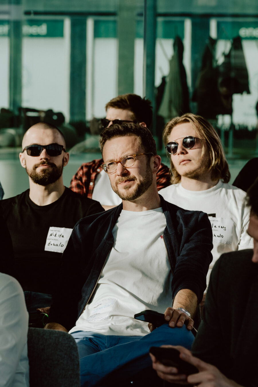 fotograf startupow rotunda reaktorX 36 Drei Männer sitzen dicht beieinander in einem sonnenbeschienenen Raum; zwei tragen Sonnenbrillen und einer eine Korrekturbrille. Sie scheinen alle aufmerksam zuzuhören und tragen Namensschilder an ihren Hemden. Im Hintergrund sind andere Menschen und große Fenster zu sehen.