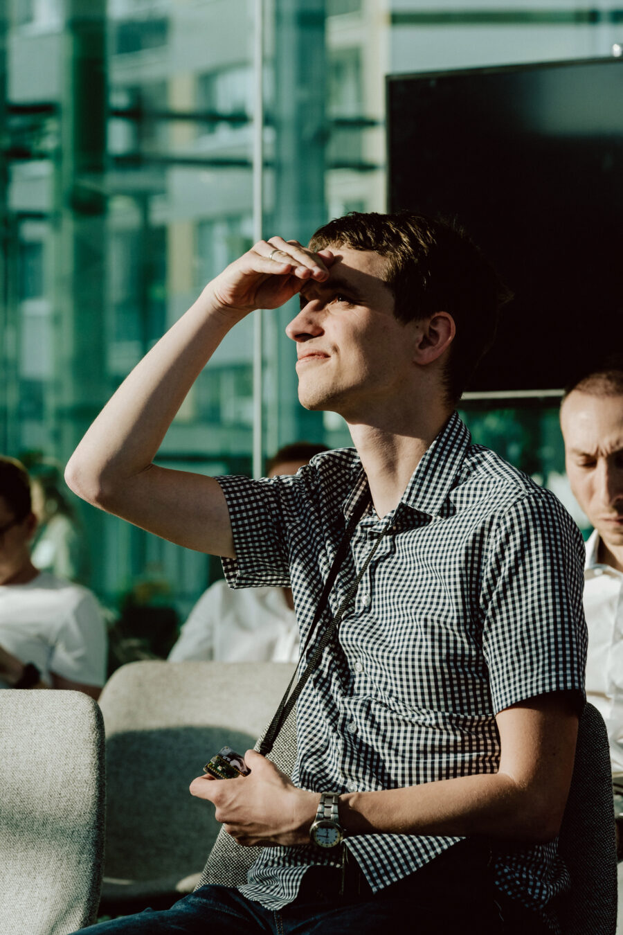 fotograf startupow rotunda reaktorX 37 Eine Person in einem karierten Hemd sitzt in einem Raum, schirmt mit einer Hand die Augen vor dem hellen Sonnenlicht ab und hält mit der anderen einen Gegenstand. Andere Menschen sitzen in der Nähe und das Sonnenlicht strömt durch große Glasfenster herein.