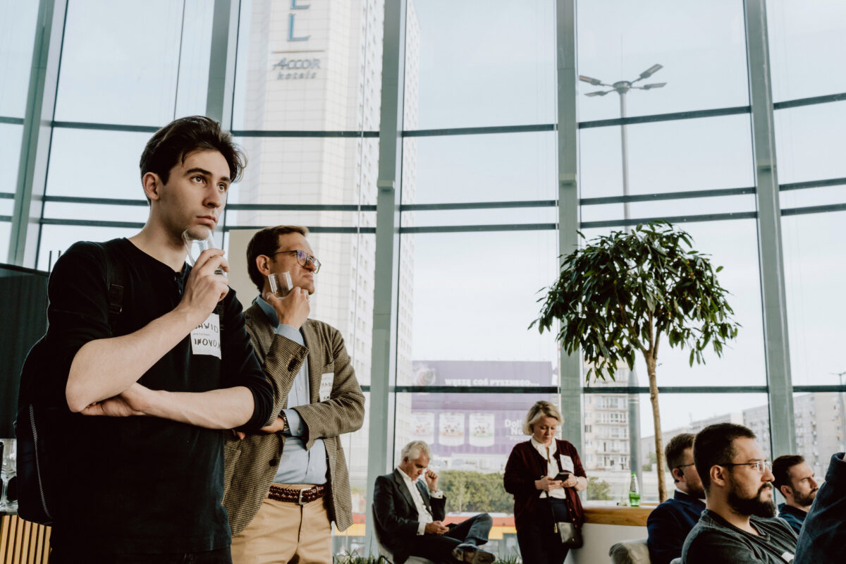 fotograf startupow rotunda reaktorX 39 Eine Gruppe von Menschen, meist Männer, sitzen und stehen in einem Raum in der Nähe großer Fenster und beobachten aufmerksam etwas im Hintergrund. Einer der Männer im Vordergrund hält sich nachdenklich das Kinn. Draußen sind die Gebäude der Stadt zu sehen.